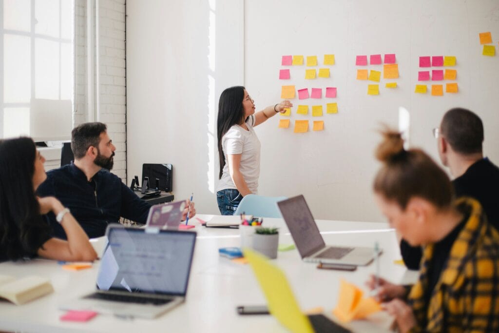 Equipe de trabalho em reunião colaborativa; uma mulher em pé organiza post-its coloridos em uma parede enquanto os colegas acompanham sentados à mesa com notebooks. Avance SEO. Marketing.